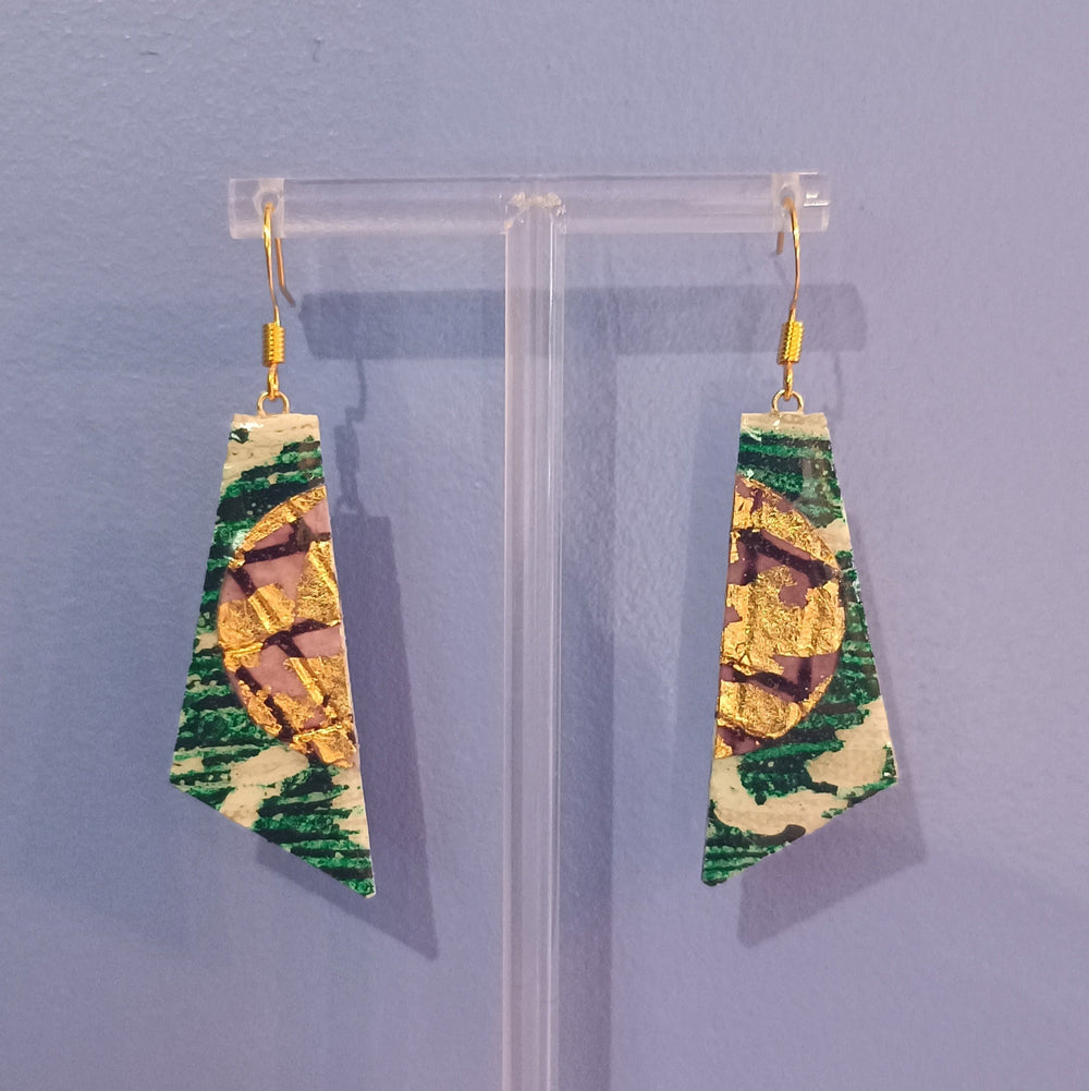 The Rothlú Cora Batik Textile Earrings in Dark Green, Gold, and Aubergine feature unique handmade patterns with gold-plated, hypo-allergenic hooks. Displayed on a clear stand against a light purple background.