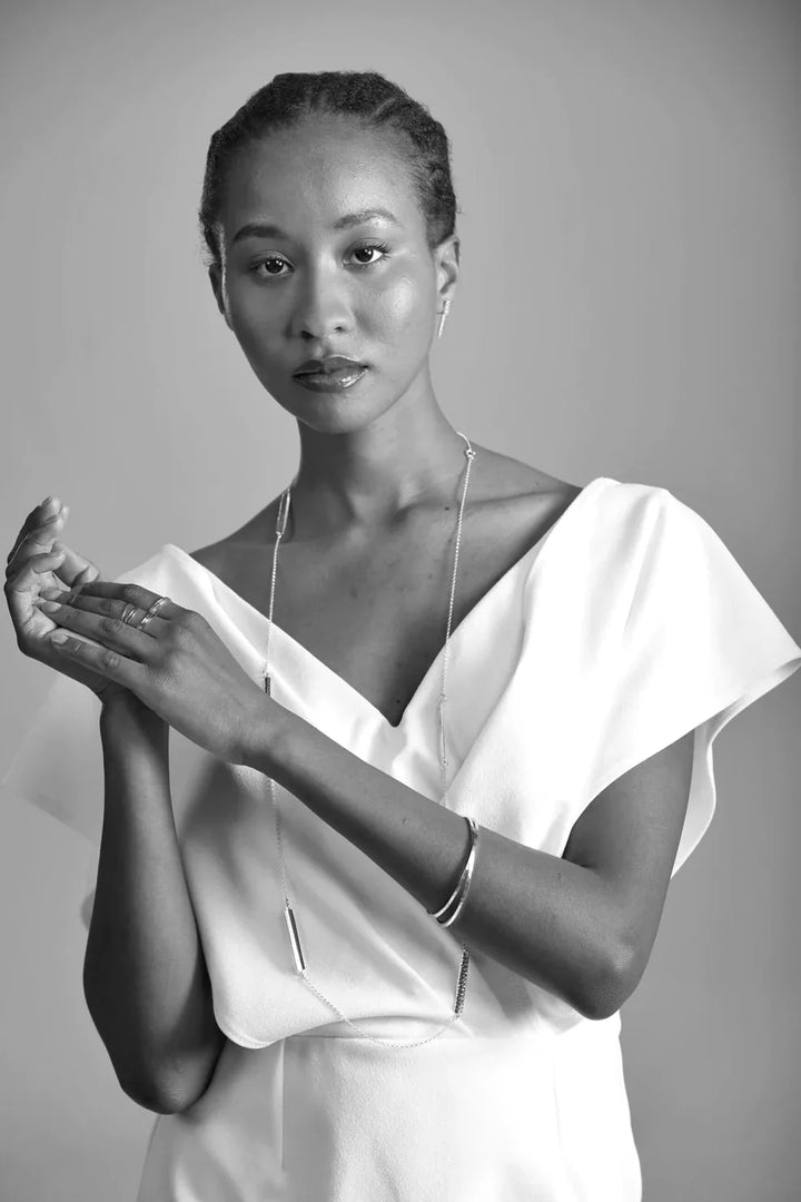 Black and white photo of a woman in a white outfit and minimalist handmade jewellery by Lynsey de Burca, including the Doon Bangle. She gazes neutrally at the camera, hair pulled back, hands gently clasped against a plain background.
