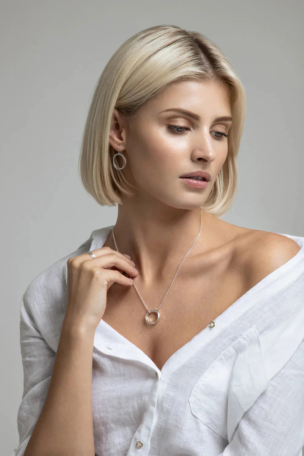 A woman with short blonde hair in a white blouse looks down, showcasing the Lynsey de Burca Doorus Silver Pendant featuring five circle links and matching earrings, complemented by her subtle, natural makeup look.