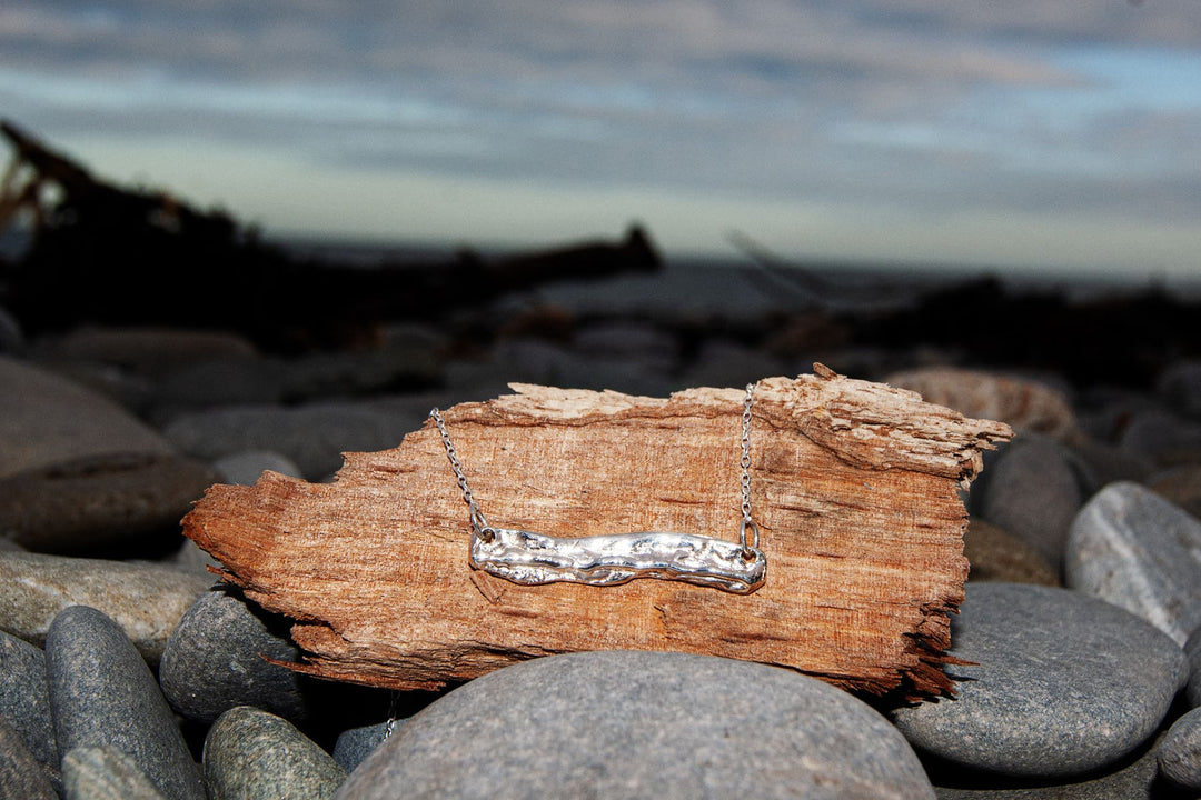 The Driftwood Silver Pendant by Ava Jane Jewellery features a textured sterling silver bar, displayed on driftwood with smooth gray stones along Dublin Bay’s rocky beach, set against a cloudy sky and soft-focus background.