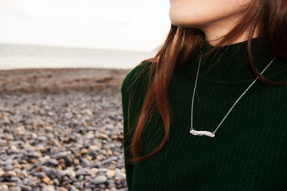 A woman with long brown hair wearing a dark green ribbed top and an Ava Jane Jewellery Driftwood Silver Pendant stands sideways on a rocky Dublin Bay beach, gazing out to sea.