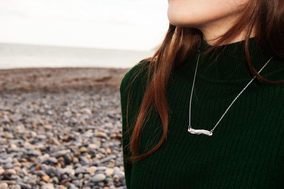 A woman with long brown hair wearing a dark green ribbed top and an Ava Jane Jewellery Driftwood Silver Pendant stands sideways on a rocky Dublin Bay beach, gazing out to sea.