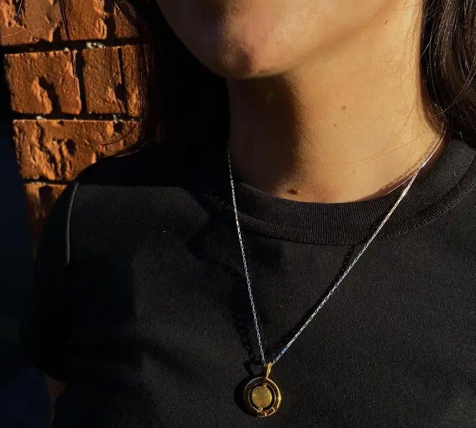 A person wearing a black shirt stands against a textured brick wall. They have a Gallardo & Blaine Designs Urú Large Pendant & Chain hanging around their neck. The sunlight casts shadows, partially illuminating their face and chest.