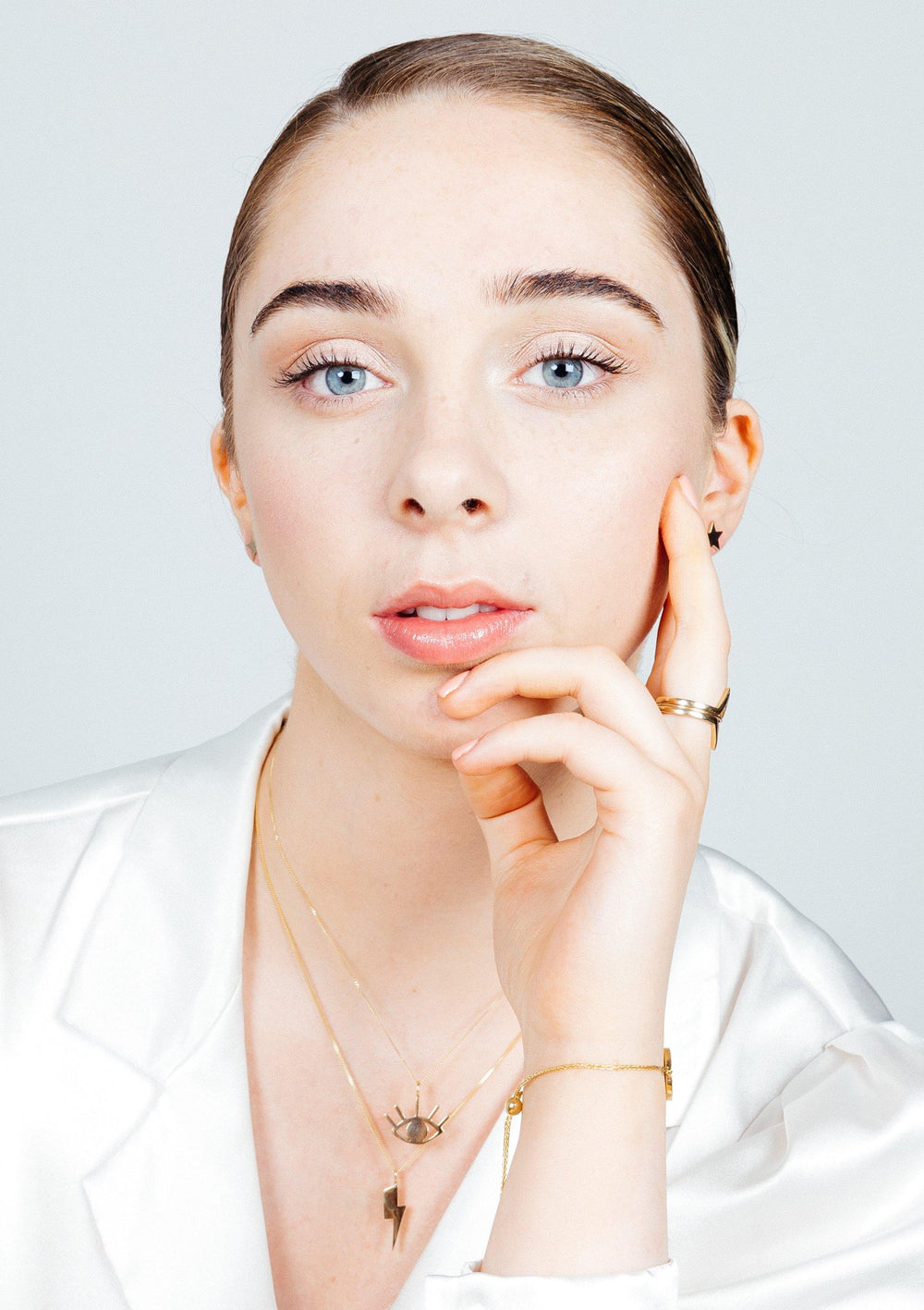 A young person with light skin, blue eyes, and brown hair pulled back gazes directly at the camera, resting their chin on their hand. They are wearing a white shirt and gold jewelry from Cosmic Boulevard, including a Bowie Lightning Bolt Pendant in 9ct Gold (available in yellow or rose gold), along with a gold ring. Their minimal makeup enhances their natural look.