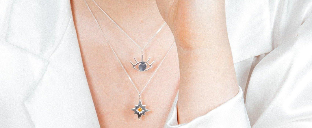 Close-up of a person wearing a white top, showcasing two layered necklaces: one with a Cosmic Boulevard Silver Evil Eye Pendant and another with a star-shaped talisman featuring an orange gemstone in the center. The person’s hand is gently touching their neck.