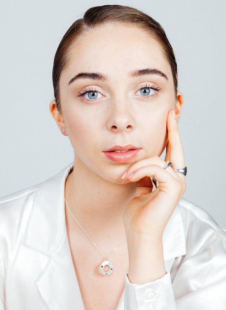 A person with light skin, blue eyes, and dark blonde hair slicked back is wearing a white silky top and a Cosmic Boulevard Shooting Star Rainbow Pendant Silver. They rest their chin on one hand while the other hand touches their face, looking directly at the camera. The background is light gray.