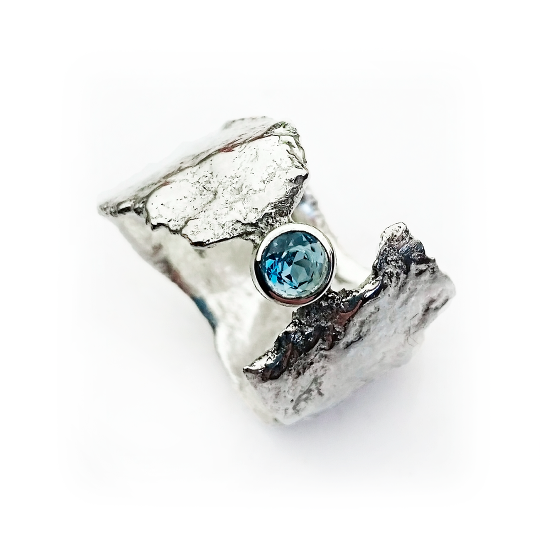 A rugged, silver ring with a textured, uneven surface featuring a prominent, round Blue Topaz gemstone set in the center. The design is organic and rough, giving the Faerie Tale Ring In Blue Topaz by Caroline Stokesberry-lee an ethereal faerie tale appearance.