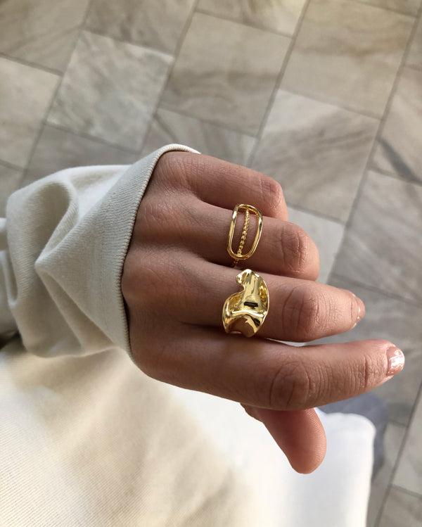 Close-up of a hand wearing two Bonvo Flor Rings—one gold plated, one sterling silver—with a cream long sleeve partially covering the fingers. The background features light-colored tiled flooring.