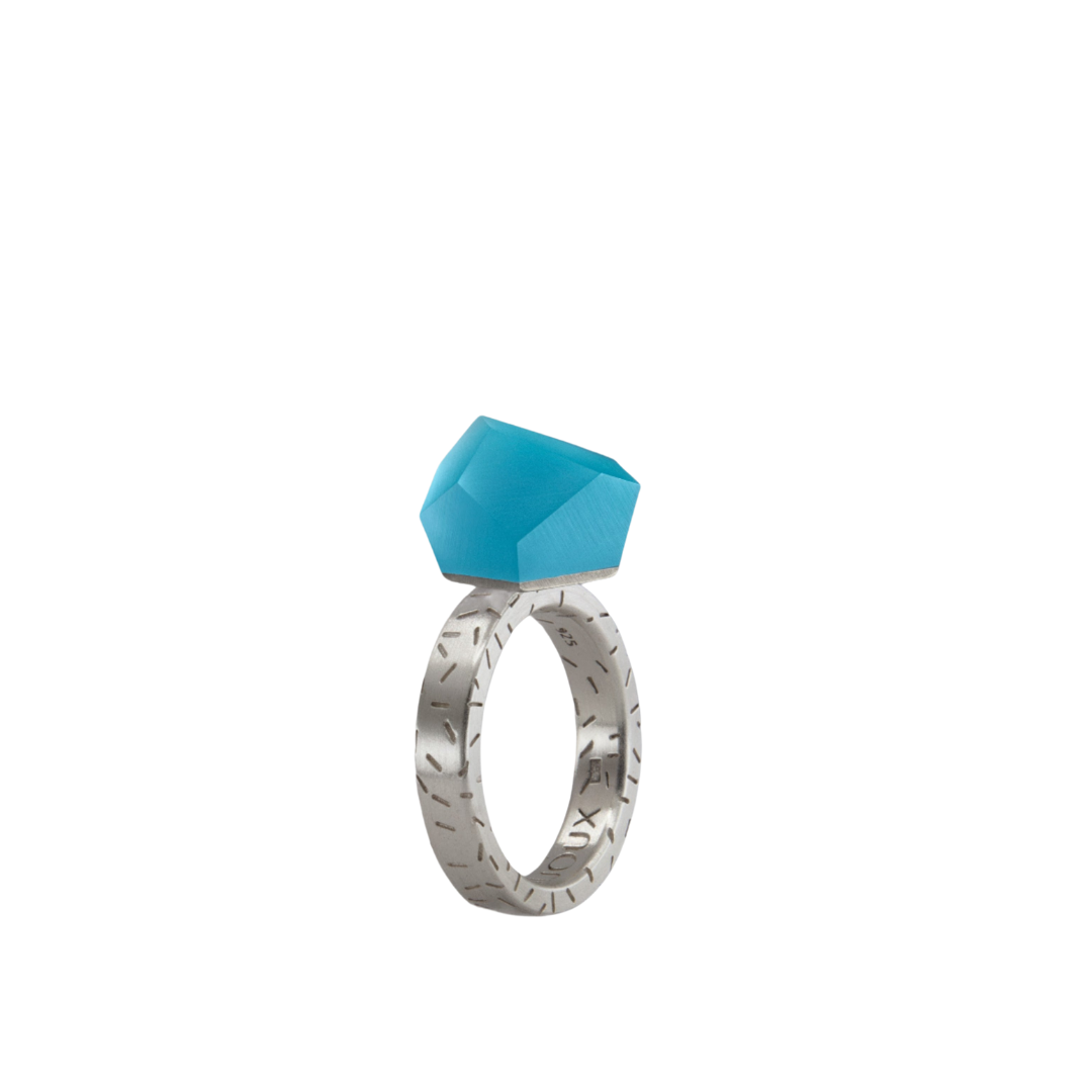 The Fruit Bijoux Ring Top VU Crystals Capri Blue by Fruit Bijoux features a modern, minimalist sterling silver band with engraved patterns and a large faceted geometric blue stone, all shown against a white background.