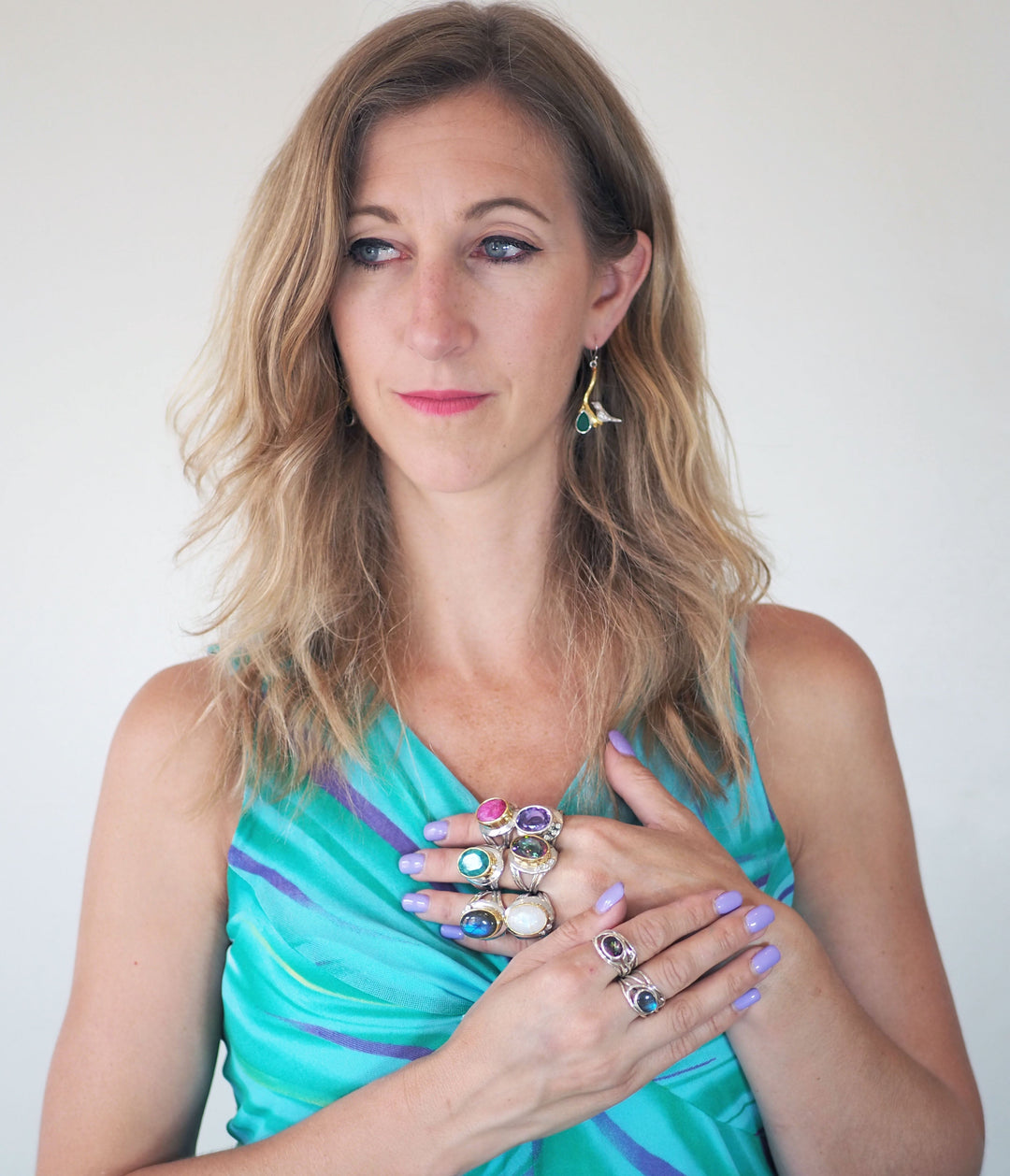 A woman with wavy blonde hair, wearing a blue-green dress and lavender nails, poses with her hands on her chest to showcase the Eyetelia Ring in various gemstones by Gallardo & Blaine Designs. She looks slightly to the side.