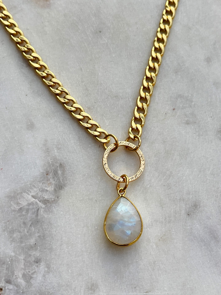 The Gaia Moonstone Necklace by Kybalion Jewellery features a gold plated curb chain with a textured circular link and an iridescent teardrop moonstone pendant, elegantly displayed on light marble.