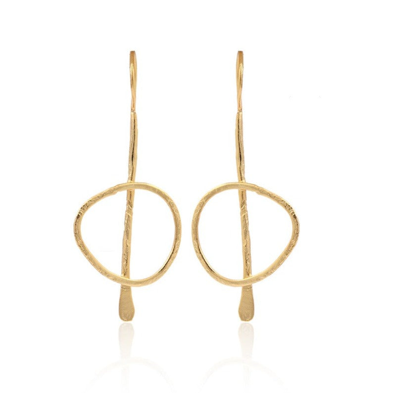 Gold plated, statement, drop earrings. The earring starts with a thin, slightly curved hook that loops through the earlobe. A slender vertical metal bar extends downward from the hook, with a slightly uneven, hand-forged texture. Attached to the vertical bar is a loosely shaped, slightly irregular circle. This frame has a hammered texture.