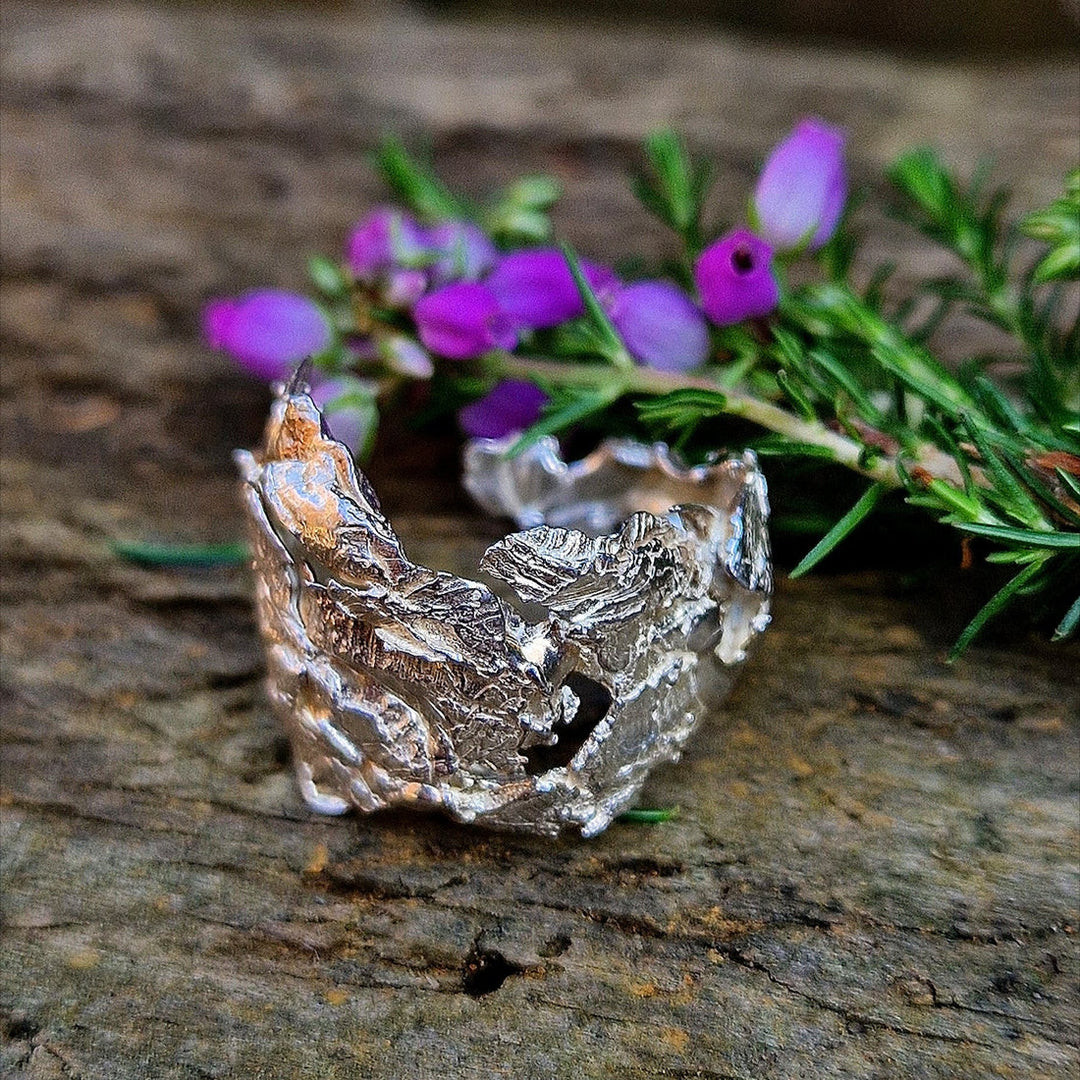 The Caroline Stokesberry-lee Goddess Of The Sea Ring, featuring a textured silver design inspired by real shells, is shown on wood with purple flowers and greenery, capturing the wild beauty of Ireland’s North Coast.