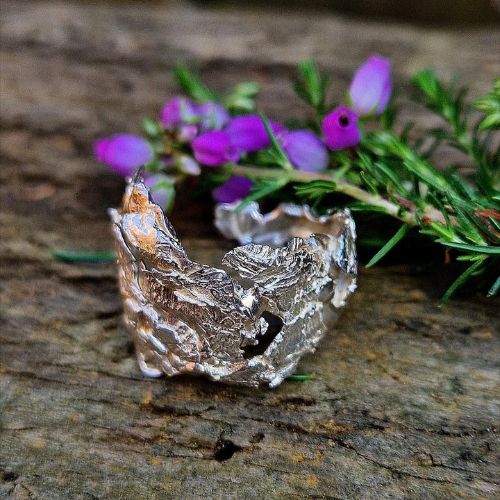 The Caroline Stokesberry-lee Goddess Of The Sea Ring, featuring a textured silver design inspired by real shells, is shown on wood with purple flowers and greenery, capturing the wild beauty of Ireland’s North Coast.