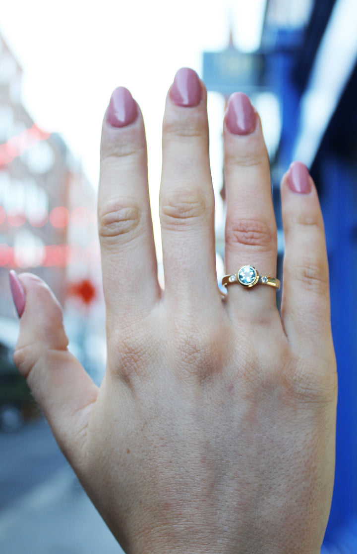 A left hand with pink nails, fingers spread, wears the Madeleine Timeless Cascade 9ct Yellow Ring on the ring finger. The background features a blurred city street.
