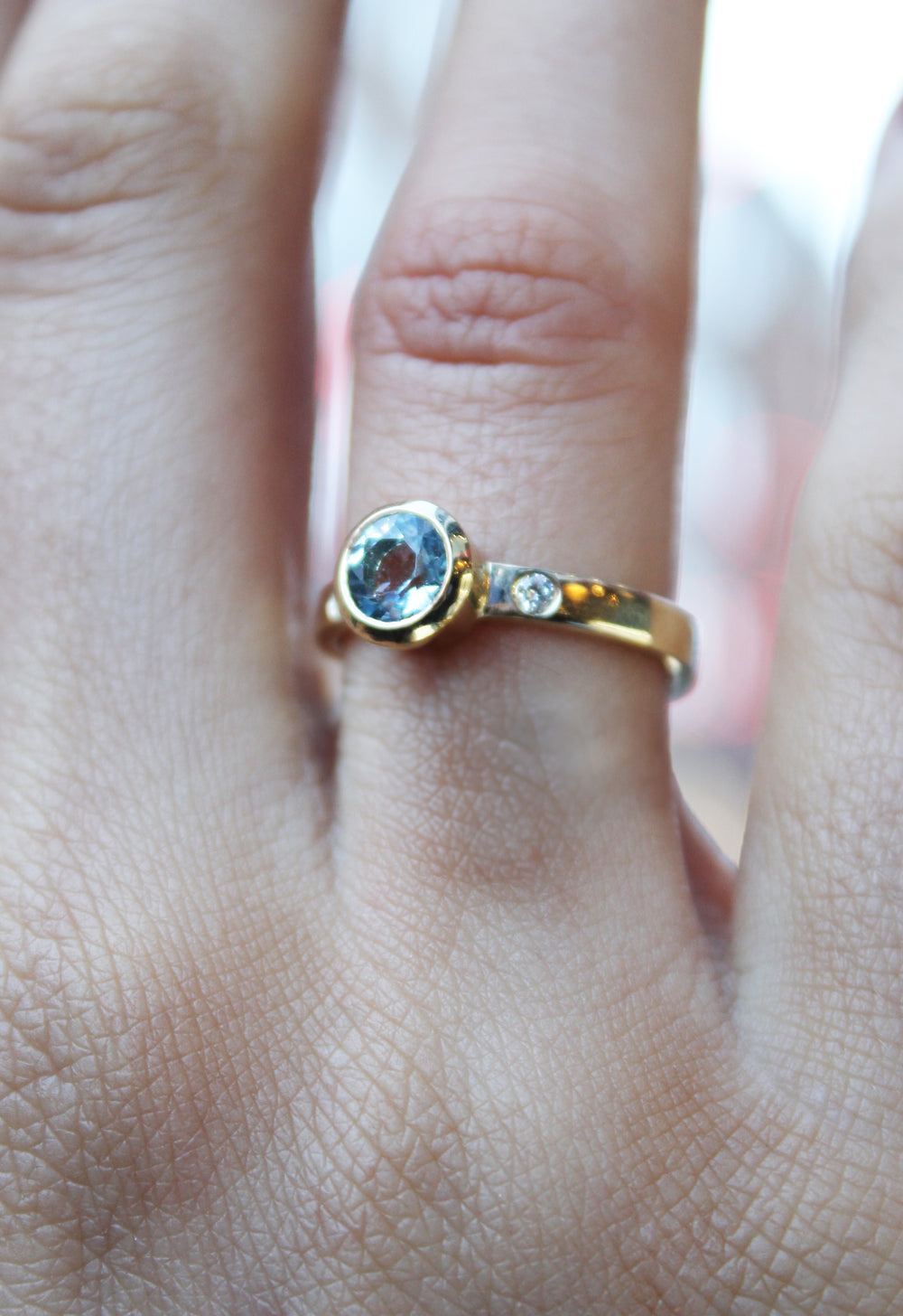 A close-up of a hand wearing Madeleine's Timeless Cascade 9ct Yellow Ring, featuring a round light blue aquamarine set in yellow gold and accented by a small clear lab grown diamond.
