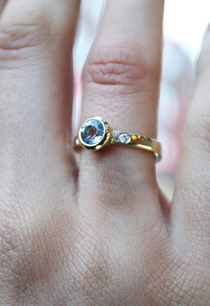 A close-up of a hand wearing Madeleine's Timeless Cascade 9ct Yellow Ring, featuring a round light blue aquamarine set in yellow gold and accented by a small clear lab grown diamond.