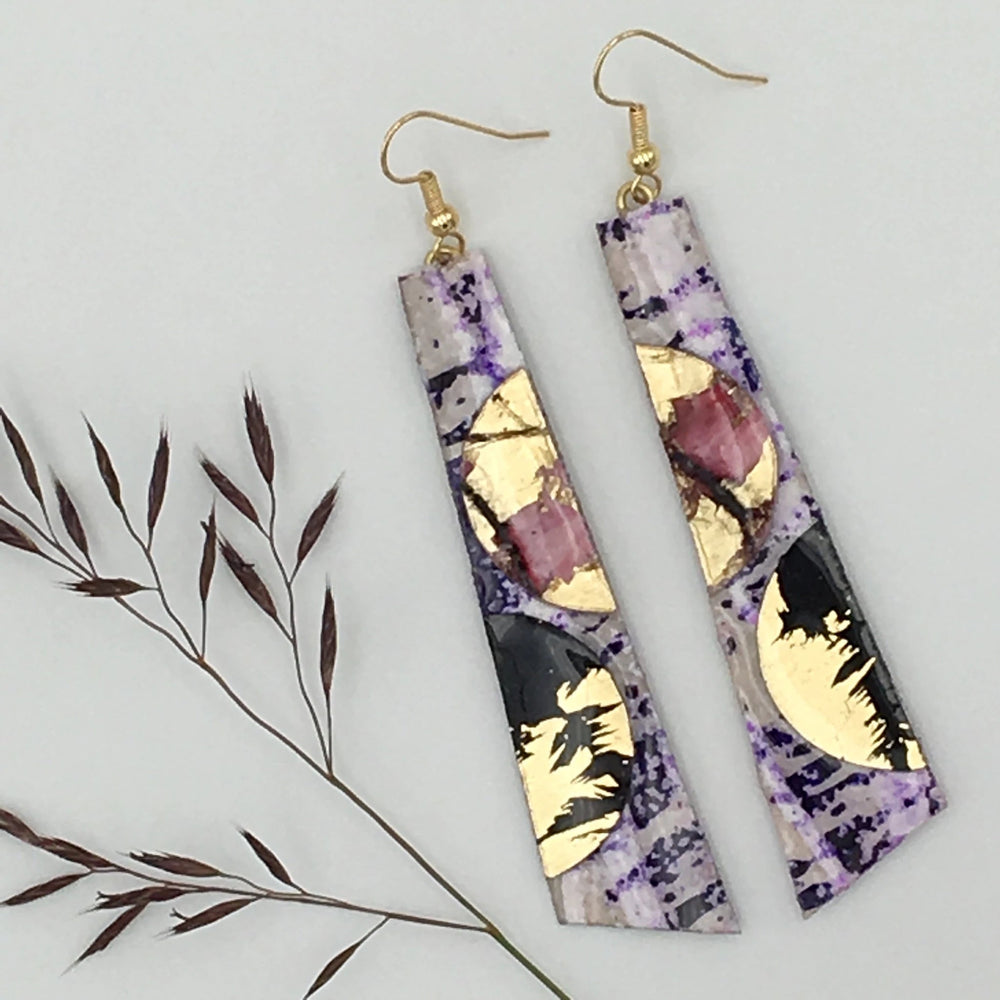 A pair of Hoop-La Batik Textile Earrings in Purple/Red/Black/Gold by Rothlú is displayed against a white background. The rectangular earrings feature an abstract pattern and have gold hooks, showcasing a unique texture that captures attention. Positioned beside them is a sprig of dried grass, enhancing their delicate charm.