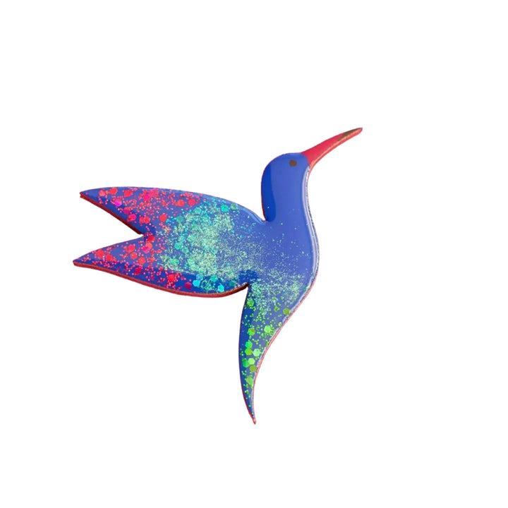 A small, vibrantly colored Hummingbird Pin Brooch by Naoi with a predominantly blue body. Its wings and tail are adorned with splashes of green, red, and yellow paint. The beak is bright red, and a small black dot represents the eye. Hand painted on wood, it stands out beautifully against the white background.
