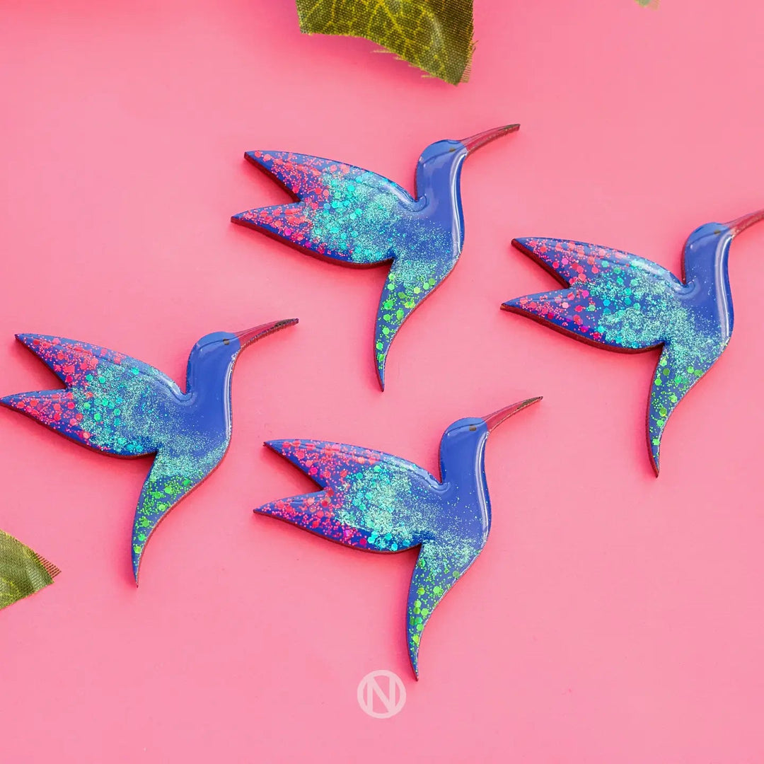 A photo showing four blue and glittery, hand-painted wooden Naoi Hummingbird Pin Brooches arranged on a pink background. The brooches have a shiny finish with varying patterns of green, blue, and pink glitter on their wings. A small portion of green leaves is visible at the top corners.