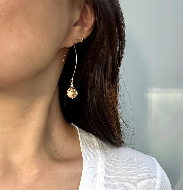 Close-up of a woman with long brown hair in a white top, wearing Aria V’s Gold Sphere Drops—metallic gold hoop earrings with textured circular pendants—set against a plain light background.