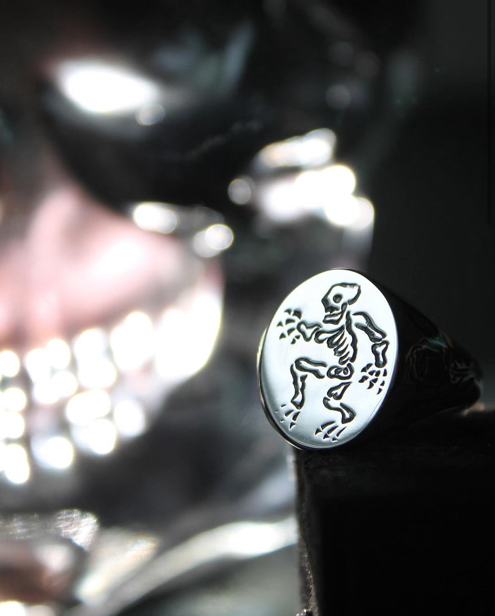 The Cosmic Boulevard Sterling Silver Memento Mori Signet Ring, engraved with a dancing skeleton, is showcased in focus on a black stand, with a blurred metallic skull seen in the background.