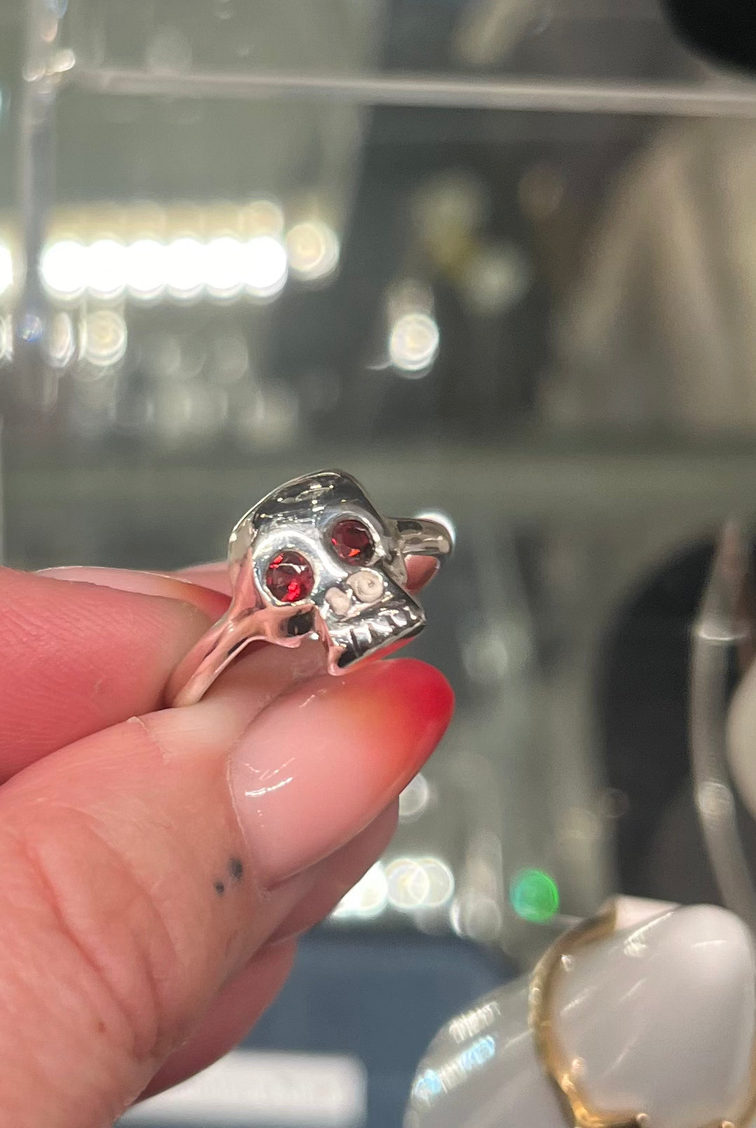 A hand with red and nude ombre nails holds the Cosmic Boulevard Silver Memento Mori Skull Ring, which features intricate teeth details and red gemstone eyes. A blurred glass display case is visible in the background.