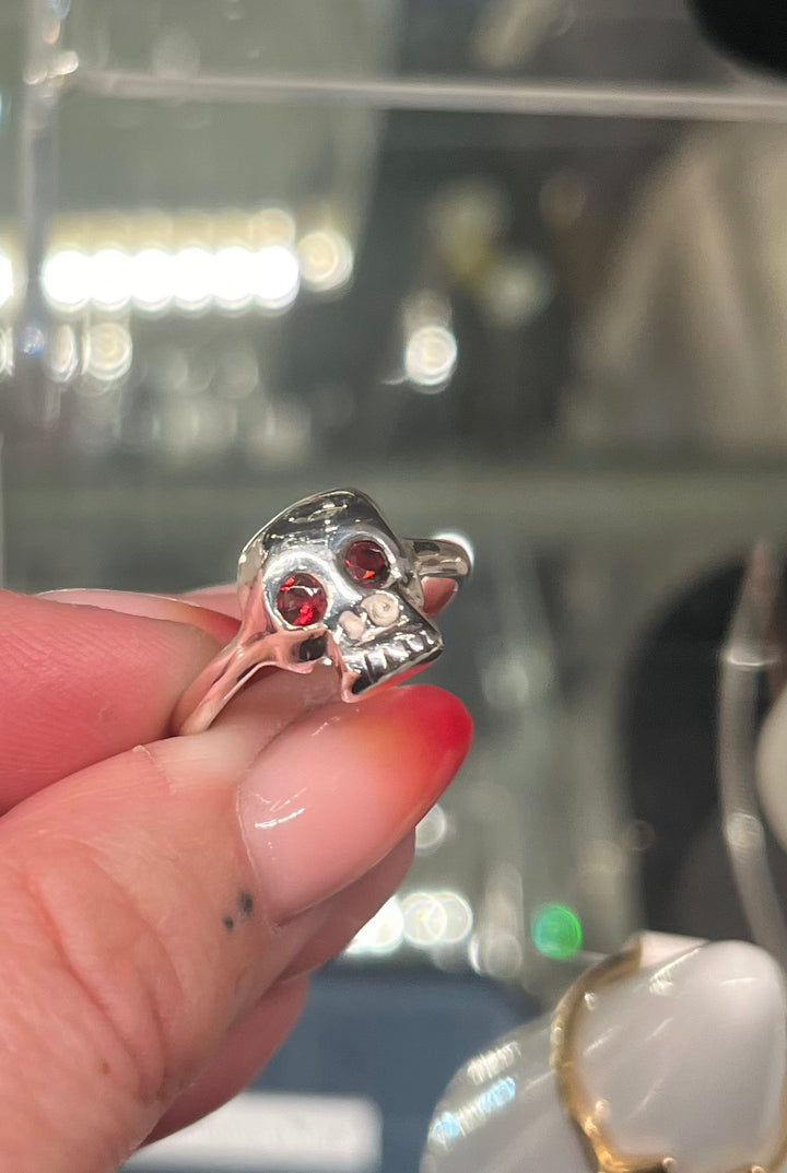 A hand with red and nude ombre nails holds the Cosmic Boulevard Silver Memento Mori Skull Ring, which features intricate teeth details and red gemstone eyes. A blurred glass display case is visible in the background.