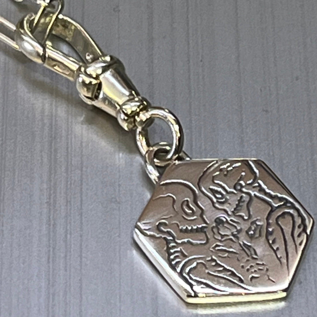 The Lovers Silver Pendant by Cosmic Boulevard features a hexagonal silver charm on a clasp, engraved with a skeleton and person face to face in a loving moment, the skeleton holding a rose.
