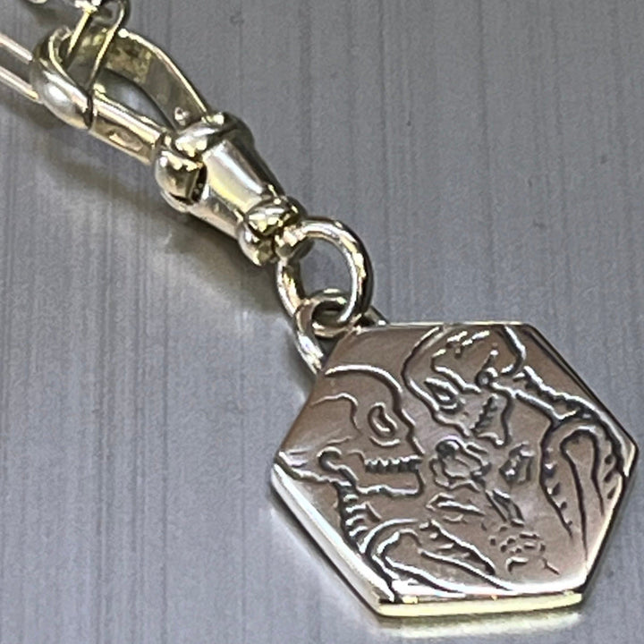 The Lovers Silver Pendant by Cosmic Boulevard features a hexagonal silver charm on a clasp, engraved with a skeleton and person face to face in a loving moment, the skeleton holding a rose.
