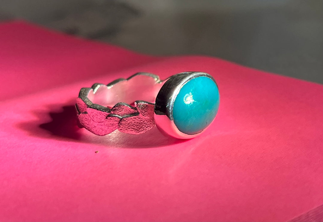 A Leoma Drew Frosted Silver ring with Turquoise featuring a bright turquoise stone set in a bezel setting. The 6mm D shape band has a textured, uneven design. The ring is placed on a piece of pink paper, creating a vibrant contrast with the turquoise stone.
