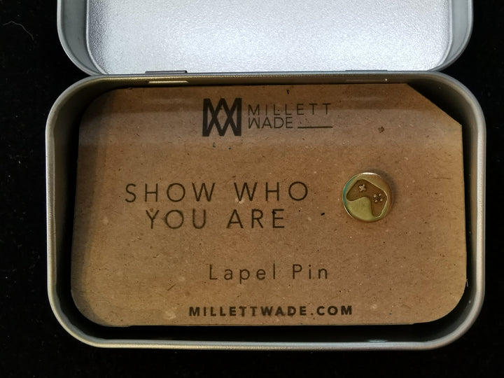 A small custom Gamer Lapel Pin rests on a card inside a metal tin. The card displays "Gamer Lapel Pin," the brand "Millett Wade," and the website "millettwade.com." Ideal for collectors of icon pins.