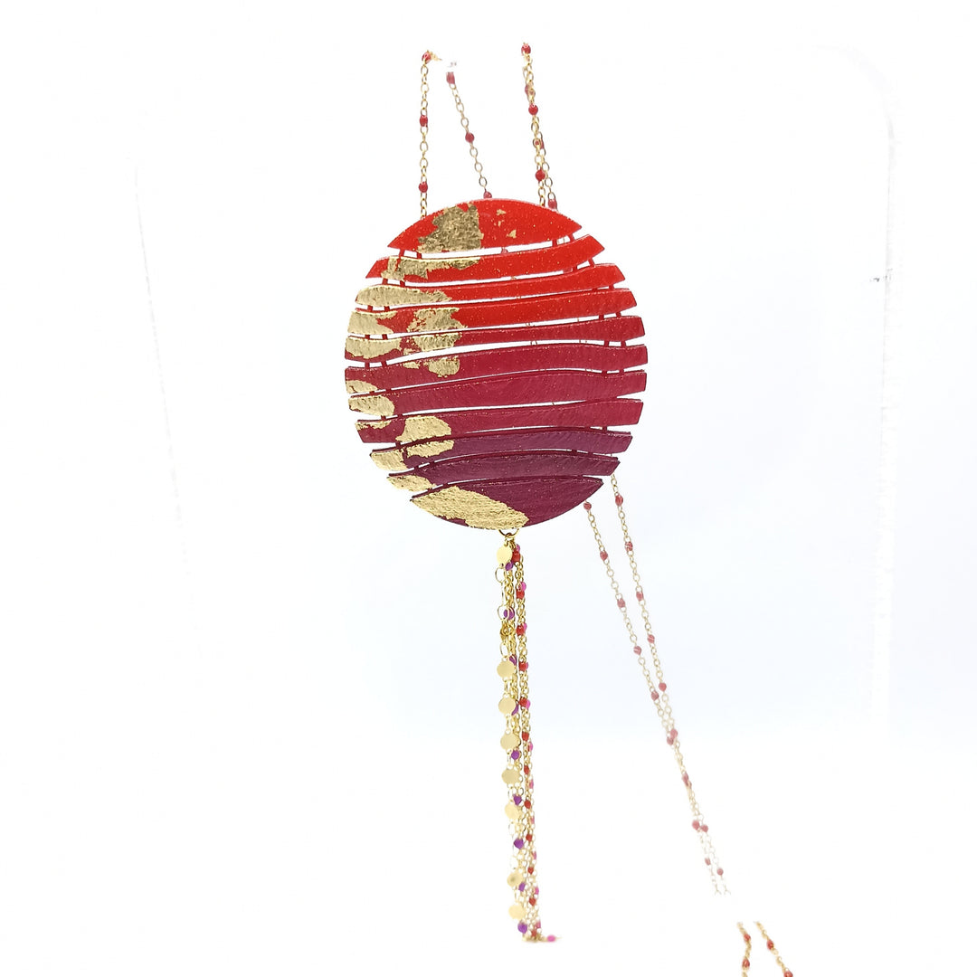 A Long Circle Wave Necklace in Red & Burgundy from Tochu-De dangles from a thin, elegant chain. The design has an artistic, modern appearance, with a small, multi-colored beaded tail extending from the bottom of the pendant. This beautiful piece boasts a 28" chain length for perfect drape.