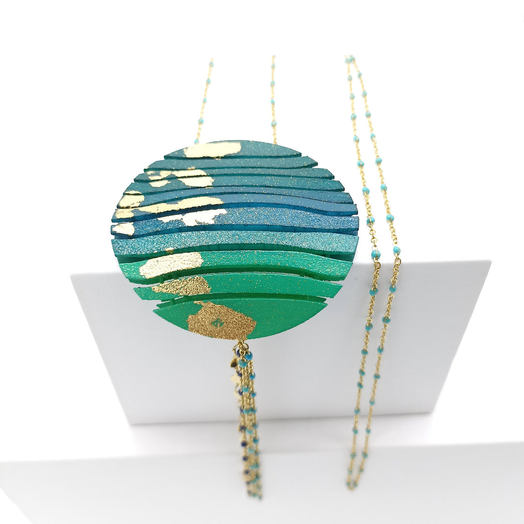 The Long Circle Wave Necklace in Petrol & Emerald by Tochu-De features a round pendant with wavy green, blue, and gold patterns on a delicate beaded chain, shown against a white background with a folded card behind it.