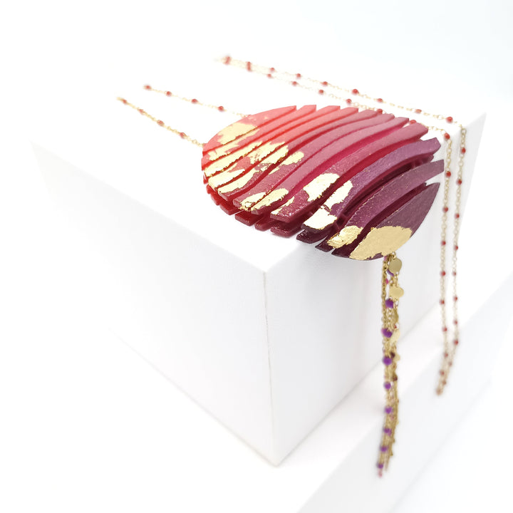 The Long Circle Wave Necklace in Red & Burgundy by Tochu-De features sculptural layered curves with gold accents, hanging from a 28-inch beaded chain, and is draped over a white geometric block for display.