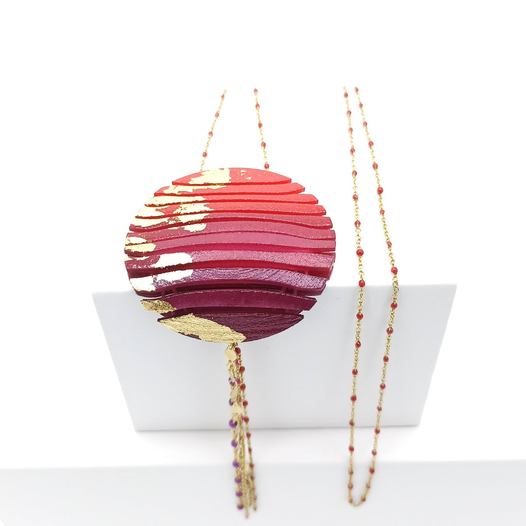 A circular red pendant with gold accents hangs from a delicate, beaded chain. The pendant features a layered, textured design, creating a striking and elegant look. The Tochu-De Long Circle Wave Necklace in Red & Burgundy is 3D printed in nylon and displayed against a simple white background.