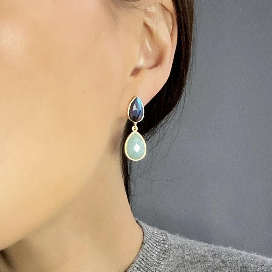 A close-up of a person wearing Aria V’s Blue Lagoon Earrings—gold brass, handmade in Ireland, with a dangling design featuring a small dark blue teardrop above a larger light green teardrop.