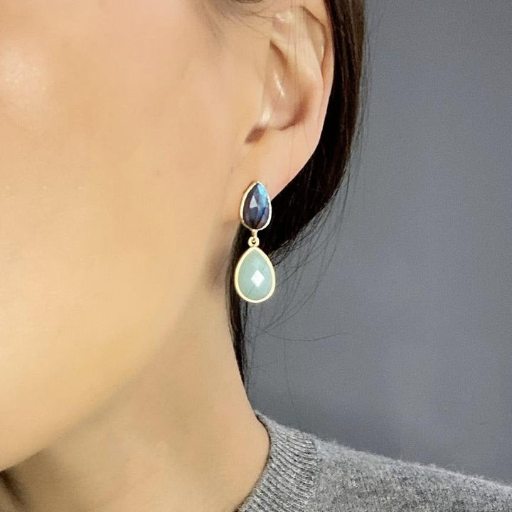 A close-up of a person wearing Aria V’s Blue Lagoon Earrings—gold brass, handmade in Ireland, with a dangling design featuring a small dark blue teardrop above a larger light green teardrop.