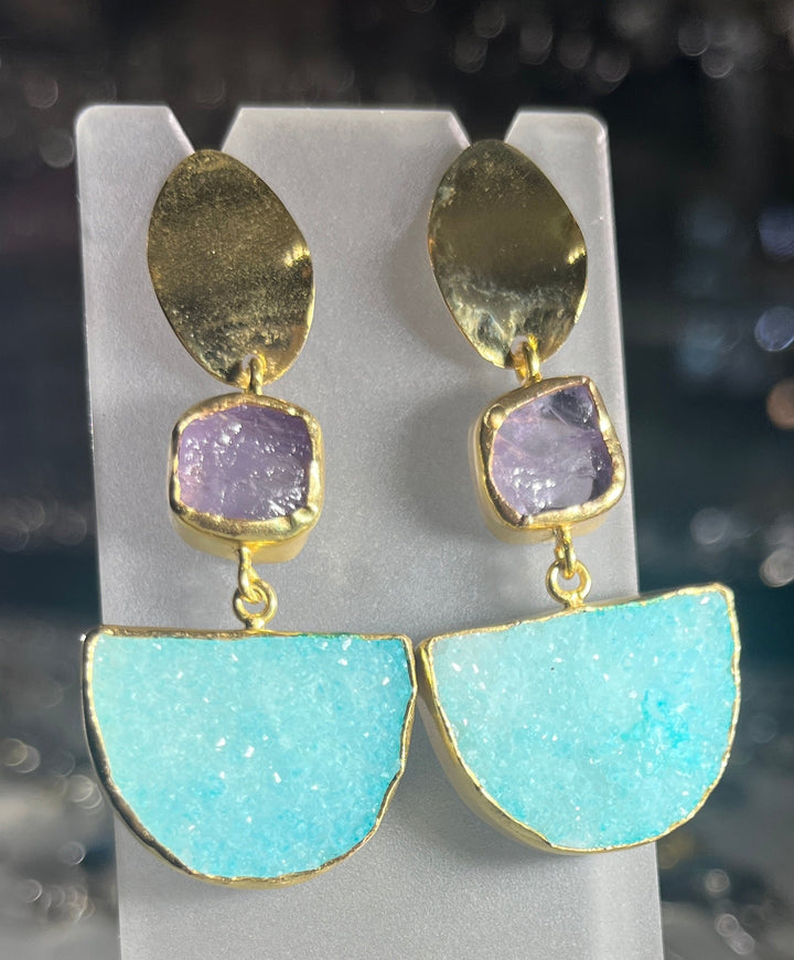 A pair of Watermelon Tropical's Amazonia Earrings In Amethyst & Aqua Druzy displayed on a stand. Each earring features a gold-toned metallic oval stud, a small square purple gemstone, and a larger semi-circle turquoise stone with a rough, sparkly texture, all connected in a vertical arrangement.
