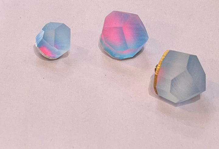 Three small, faceted, multi-colored beads with a pink and blue iridescent sheen are placed on a flat, white surface. The beads vary in size and shape, with two appearing larger and one smaller. Their unique reflective properties make them perfect for accenting the Fruit Bijoux Fruit Bijoux Ring Top Aura.