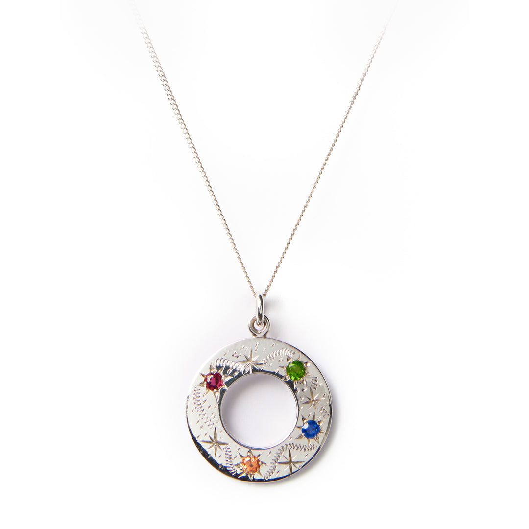 The Cosmic Boulevard Shooting Star Rainbow Pendant Silver is a sterling silver circular pendant adorned with small, colored gemstones (red, green, blue, and orange) suspended from a delicate silver chain. Handmade in Dublin, the pendant features intricate engravings and an open center, creating a hollow ring design reminiscent of shooting stars. Displayed on a plain white background.