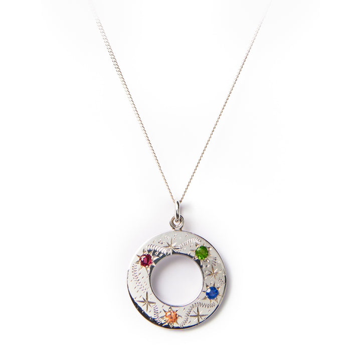 The Cosmic Boulevard Shooting Star Rainbow Pendant Silver is a sterling silver circular pendant adorned with small, colored gemstones (red, green, blue, and orange) suspended from a delicate silver chain. Handmade in Dublin, the pendant features intricate engravings and an open center, creating a hollow ring design reminiscent of shooting stars. Displayed on a plain white background.