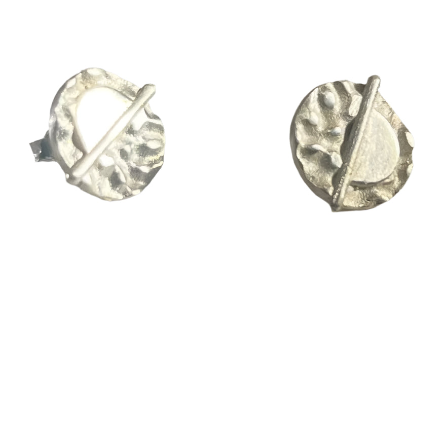 A pair of Lunar Eclipse Stud Earrings from Gallardo & Blaine Designs on a light surface. Each earring features an irregularly textured round design with an angled bar crossing the circle, creating a modern, abstract appearance reminiscent of Art Deco. The earrings are reflecting light, highlighting their shiny finish.