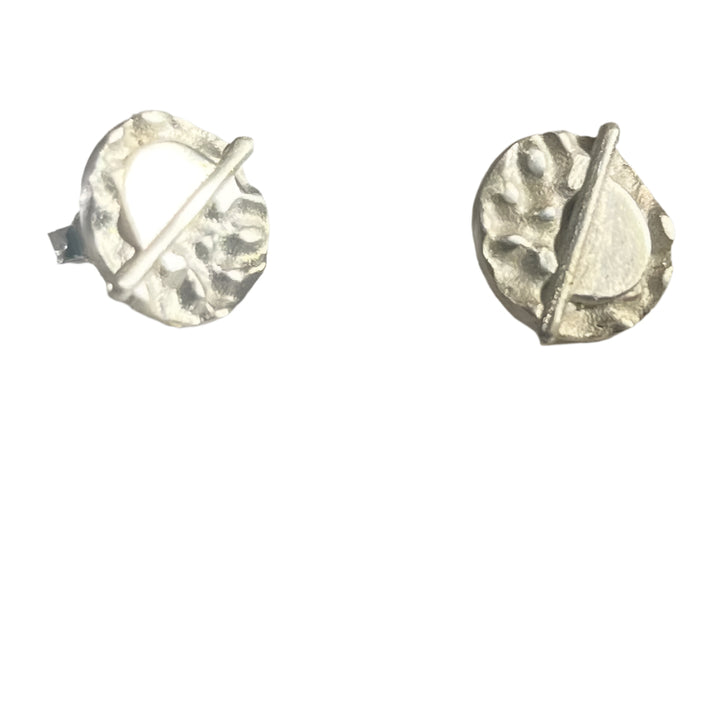 A pair of Lunar Eclipse Stud Earrings from Gallardo & Blaine Designs on a light surface. Each earring features an irregularly textured round design with an angled bar crossing the circle, creating a modern, abstract appearance reminiscent of Art Deco. The earrings are reflecting light, highlighting their shiny finish.