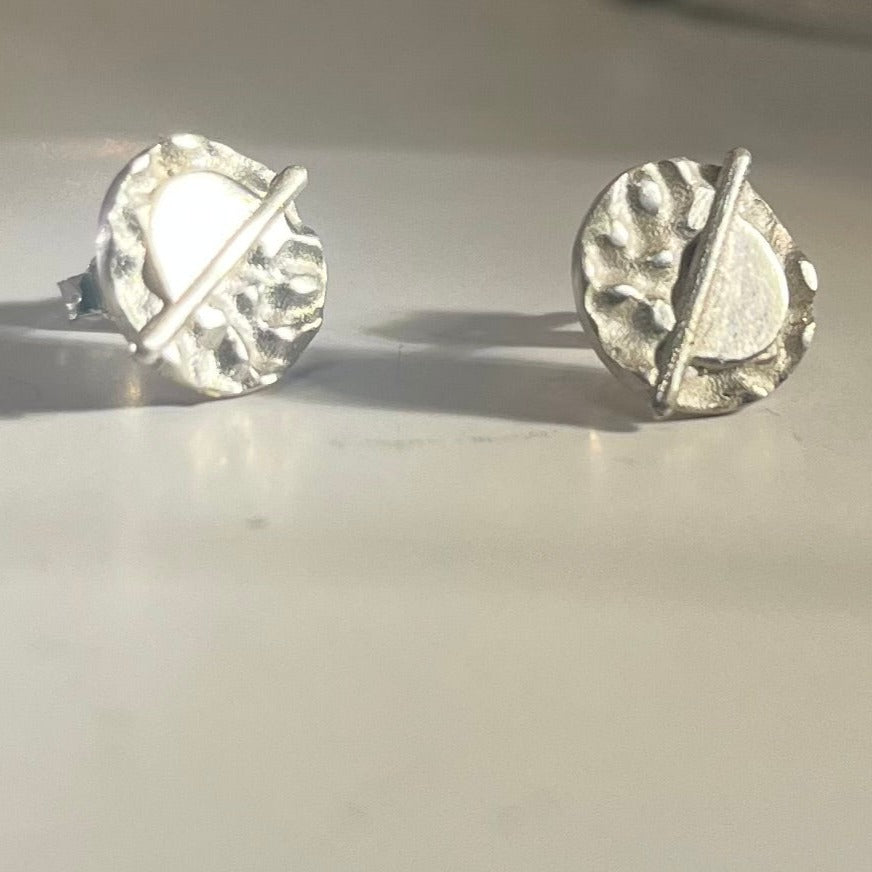 The Lunar Eclipse Stud Earrings by Gallardo & Blaine Designs feature a round, textured sterling silver design with a raised diagonal bar, shown on a smooth, light-colored surface. Also available in gold and black finishes.