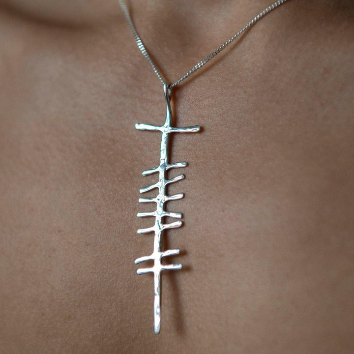 Close-up image of a silver necklace featuring a unique, abstract pendant. The Love (Grá) Large Rustic Pendant Silver by Ogham Treasure, inspired by Ogham writing, is reminiscent of a vertical line with several horizontal lines extending from one side, creating an asymmetric, ladder-like design. The background is a blurred human neckline.