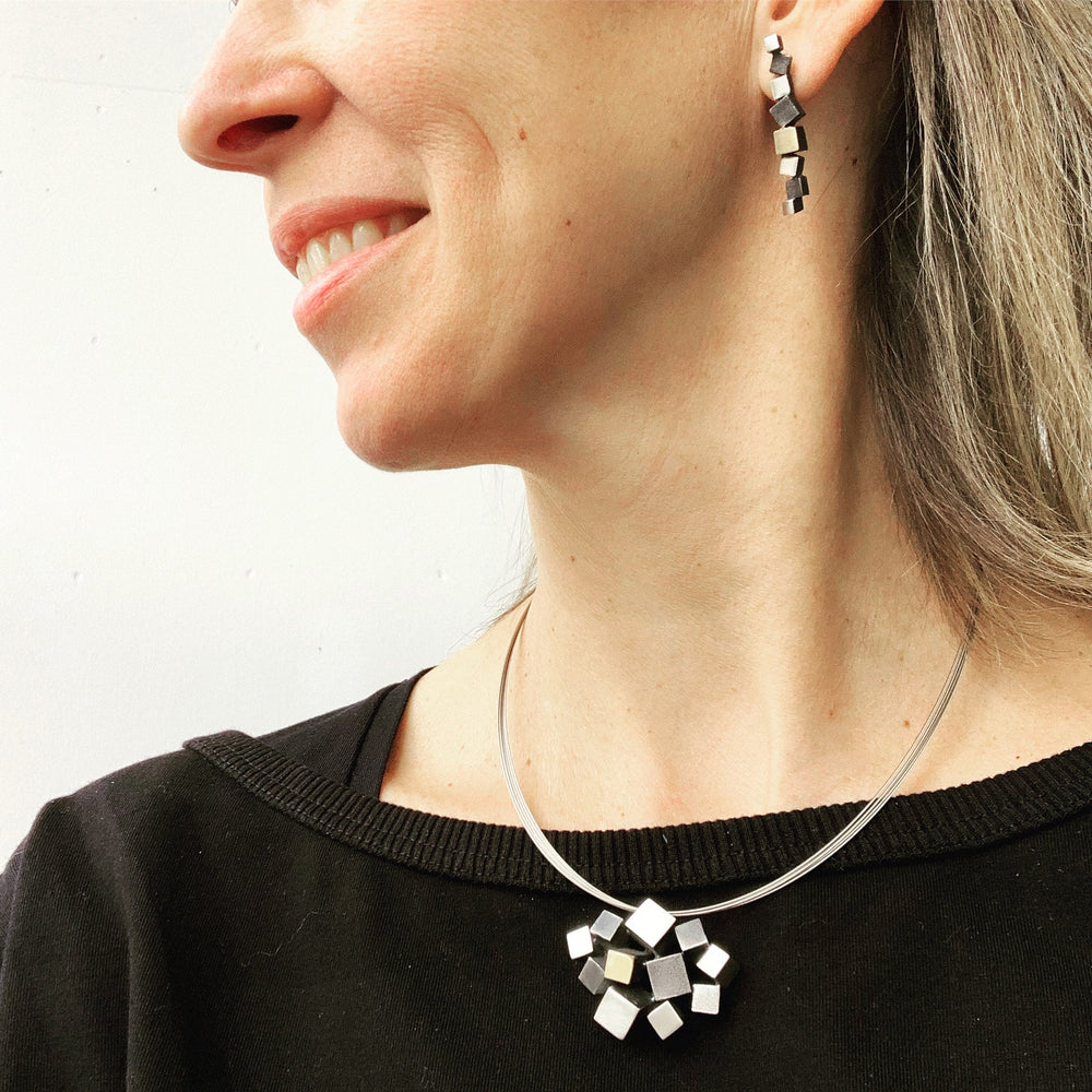 A woman wearing geometric cube earrings and an Iceland Pendant from MarCucurella smiles slightly. The handmade jewelry features a modern design with interconnected silver cubes. She is dressed in a black top.