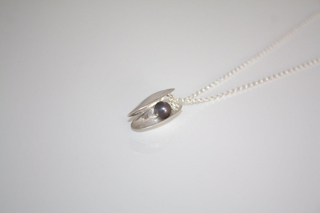 A delicate Jennifer Kinnear Open Clam Shell Necklace, available in silver or gold, is displayed on a thin chain with the locket slightly open to reveal a small freshwater pearl inside. The plain off-white background accentuates the elegance of this exquisite piece.