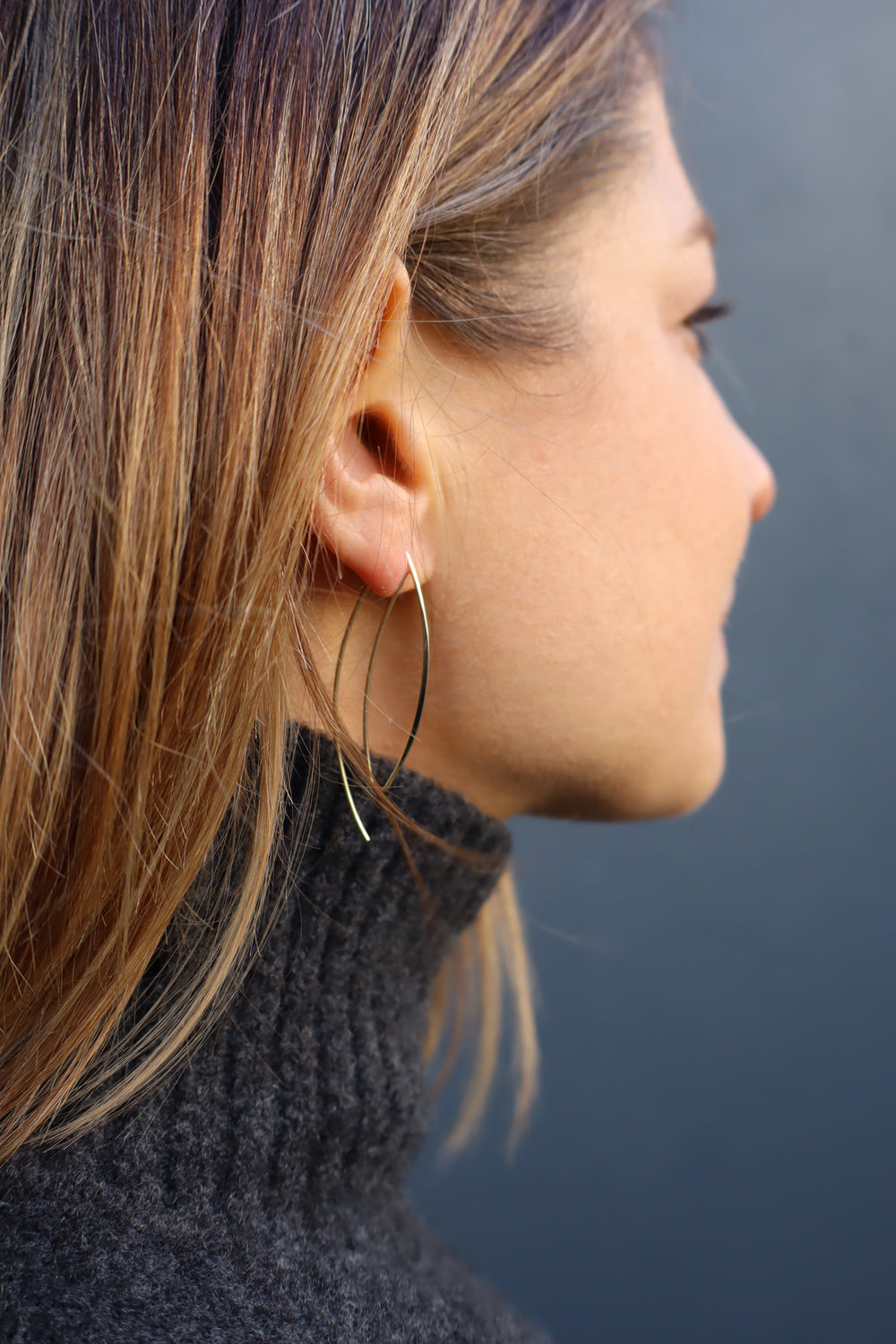 Close-up of a person with shoulder-length hair in a dark turtleneck, wearing Kybalion Jewellery's JoJo Earrings, which feature a long delicate gold-plated thread. The background is blurred and dark.