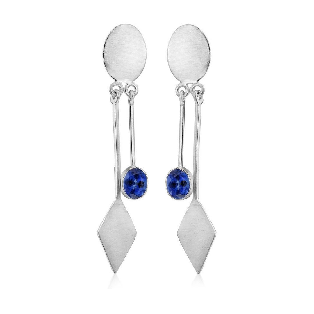 Discover modern elegance with Gallardo & Blaine Designs' Karo Earrings, silver dangle drops featuring oval studs and a sleek bar leading to a diamond-shaped drop. A small blue gemstone near the bottom enhances these chic accessories.